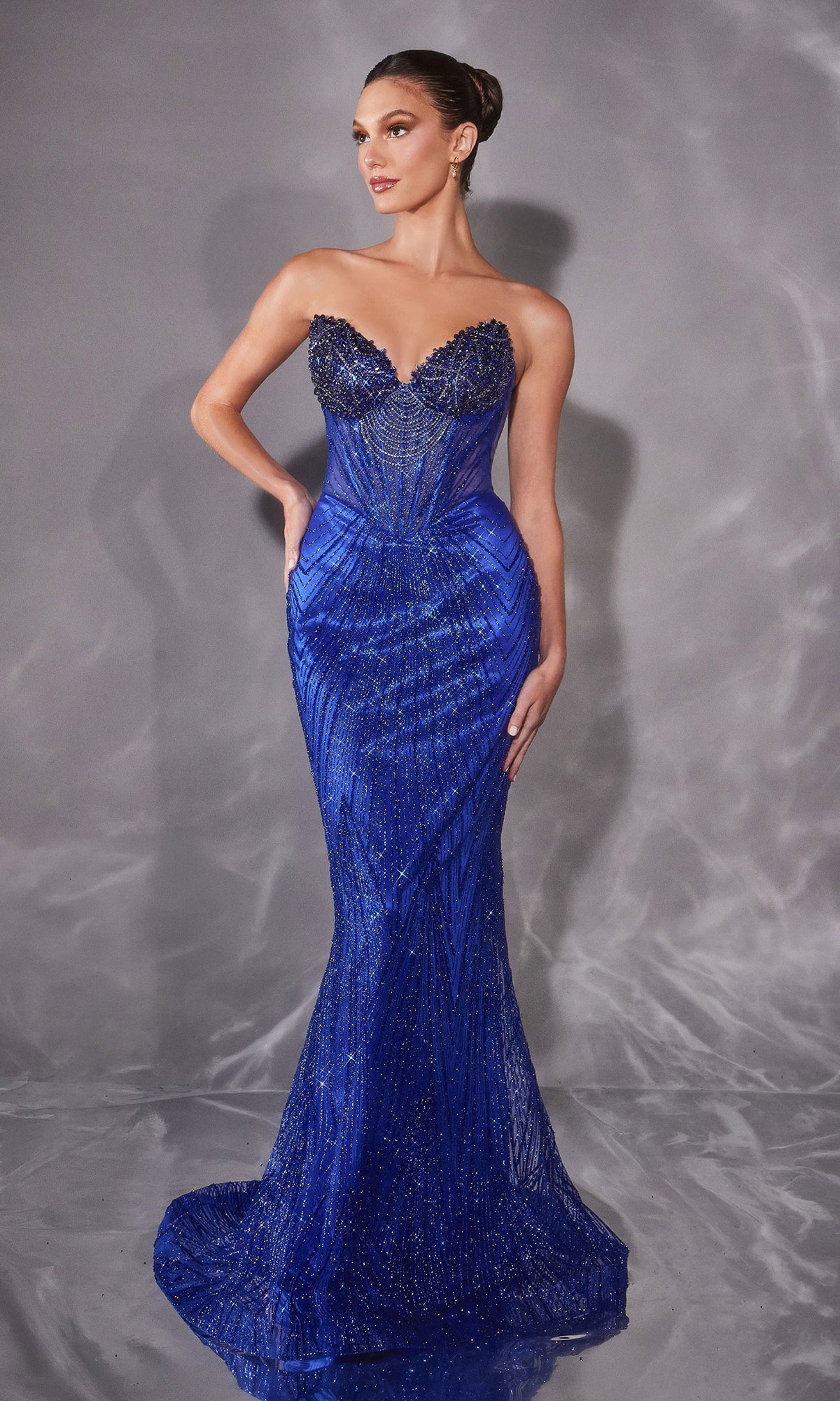 A woman in a Ladivine J903 Strapless Long Beaded Prom Dress in royal stands against a gray, textured backdrop.